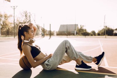 Çekici atletik genç kadın basketbol eğilerek sabah basketbol sahasında oturan açık havada poz. Moda