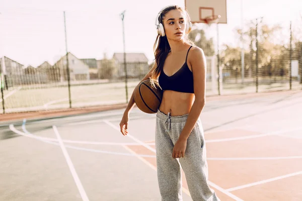 Portrait of a beautiful strong athletic woman outdoors posing while listening to music with headphones holding a basketball. — Stock Photo, Image