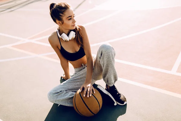 Attraktive athletische junge Frau posiert im Freien sitzend auf dem Basketballplatz und hält Basketball — Stockfoto
