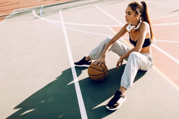 Atractiv atletic tânără femeie în aer liber pozând așezat pe teren de baschet care deține baschet — Fotografie, imagine de stoc