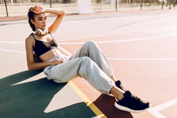 Atractiva atlética joven posando al aire libre sentada en la cancha de baloncesto por la mañana apoyada en el baloncesto. Moda — Foto de Stock