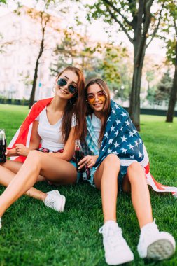 Amerikan bayrağı ile çim üzerinde Parkta oturan güneş gözlüğü iki çekici genç kız, ferahlatıcı bir içki ile