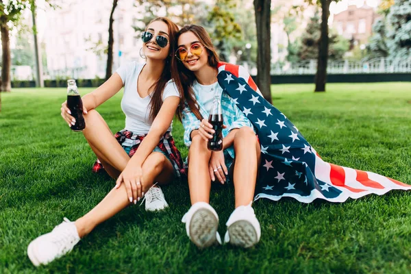 Δύο ελκυστικά νεαρά κορίτσια στα γυαλιά ηλίου που κάθονται στο πάρκο στο γκαζόν με την αμερικάνικη σημαία, με ένα δροσιστικό ποτό — Φωτογραφία Αρχείου