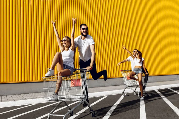 Grupa młodych ludzi w białych koszulkach, jazda zabawy na wózki w pobliżu sklepu, supermarket, przyjaciele bawią się w słonecznej pogodzie na zewnątrz — Zdjęcie stockowe
