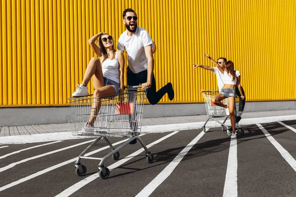 Un grup de tineri fericiți, care se distrează pe cărucioare de cumpărături, merg pe un coș de cumpărături. Ziua de vară cu lumina soarelui . — Fotografie, imagine de stoc