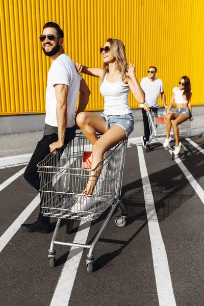Un grupo de jóvenes en camisetas blancas, paseo divertido en carritos cerca de la tienda, supermercado, amigos se divierten en el clima soleado al aire libre — Foto de Stock