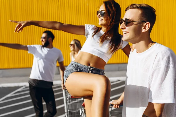 Un groupe de jeunes en t-shirts blancs, promenade amusante sur des chariots près du magasin, supermarché, amis s'amusent par temps ensoleillé en plein air — Photo