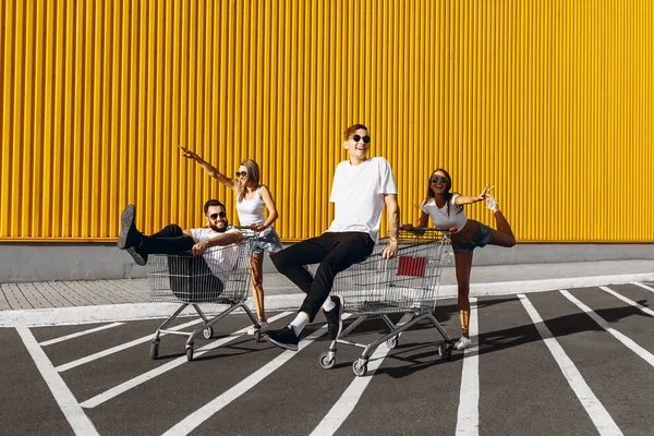 Un groupe de jeunes en t-shirts blancs, promenade amusante sur des chariots près du magasin, supermarché, amis s'amusent par temps ensoleillé en plein air — Photo