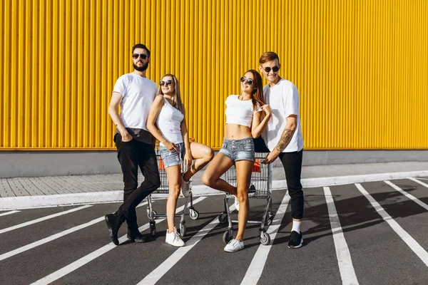 Un groupe de jeunes amis, en t-shirts blancs, s'amuser, couples posant debout près des chariots, près du supermarché — Photo