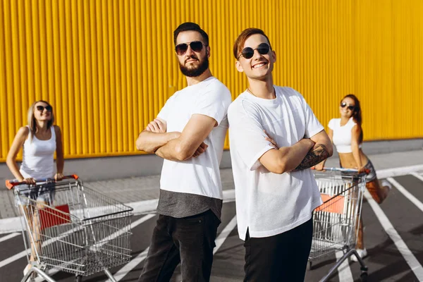 La compagnia di giovani, ragazze e ragazzi alla moda, gli amici si divertono con i carri, all'aperto vicino al supermercato nel tempo soleggiato — Foto Stock