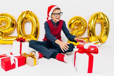 Noel Baba şapkalı mutlu çocuk, Noel hediyeli şık bebek, beyaz arka planda altın 2020 rakamlarıyla oturuyor.