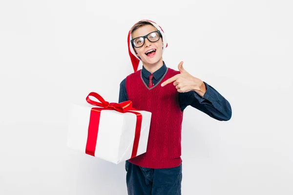 Szczęśliwy chłopak w czerwonym kapeluszu Mikołaja. Stylowy chłopiec z prezentem świątecznym, z radością wskazując palcem na prezent, na białym tle — Zdjęcie stockowe