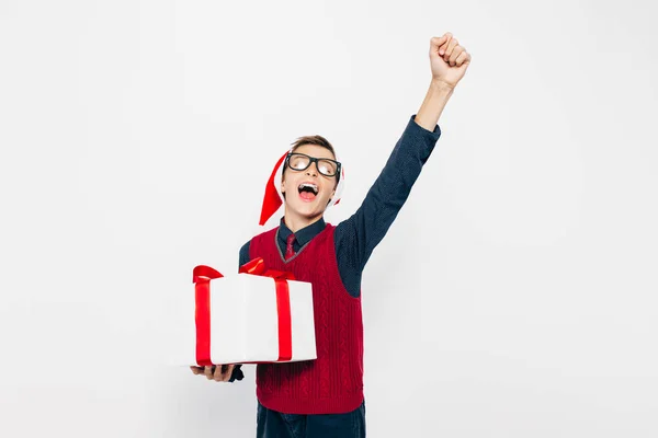 Szczęśliwy chłopak w czerwonym kapeluszu Mikołaja. Stylowy chłopiec z prezentem świątecznym zabawy i pokazując gest zwycięstwa i sukcesu na białym tle — Zdjęcie stockowe