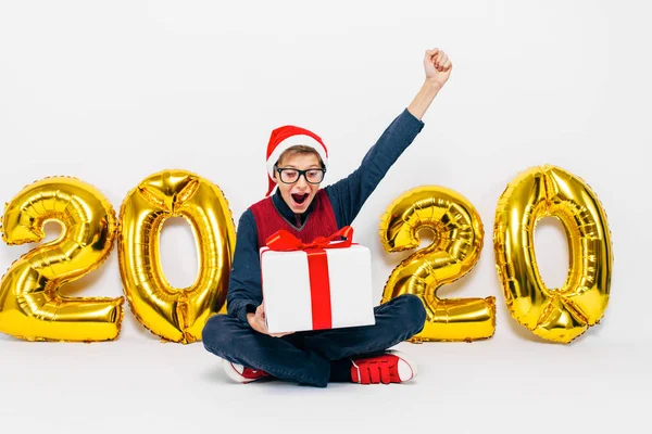 Mały chłopiec w Santa kapelusz, stylowe dziecko raduje się z prezentem świątecznym pokazując gest zwycięstwa siedząc na białym tle z 2020 złote cyfry — Zdjęcie stockowe