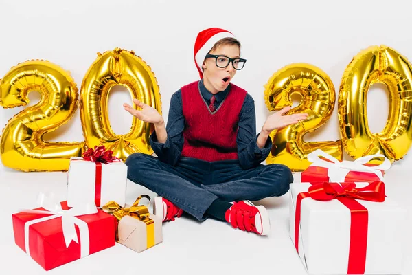 Menino feliz em chapéu de Santa, criança elegante encantado com presentes de Natal sentado no fundo branco com figuras de ouro 2020 — Fotografia de Stock