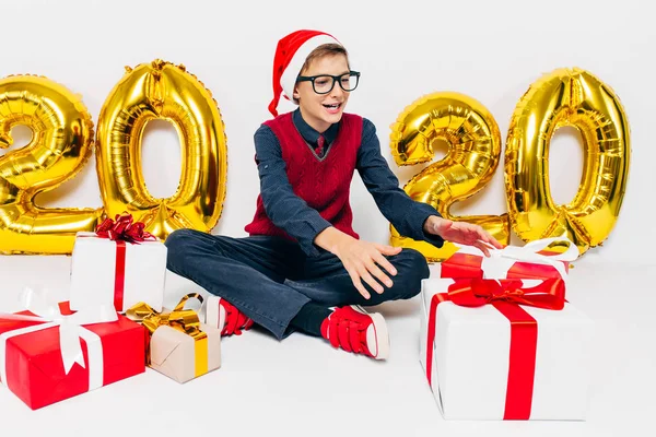 Menino feliz em chapéu de Santa, bebê elegante com presentes de Natal, senta-se no fundo branco com números de ouro 2020 — Fotografia de Stock