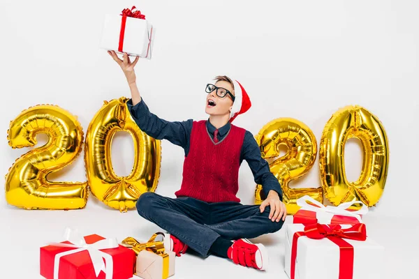 Happy kleine jongen in Santa hoed, stijlvolle baby met kerstcadeaus, zit op een witte achtergrond met goud 2020 nummers — Stockfoto