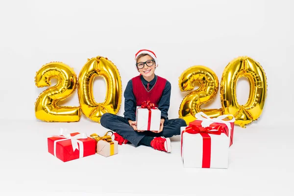 Happy little boy w Santa kapelusz, stylowe dziecko z prezentów świątecznych, siedzi na białym tle ze złotem 2020 numery — Zdjęcie stockowe