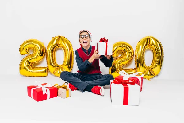 Happy little boy w Santa kapelusz, stylowe dziecko z prezentów świątecznych, siedzi na białym tle ze złotem 2020 numery — Zdjęcie stockowe