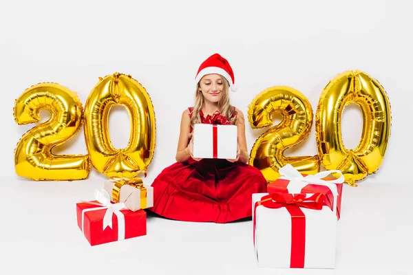 Happy little girl w Santa kapelusz, stylowe dziecko siedzi z prezentów świątecznych siedzi na białym tle ze złotem 2020 numery — Zdjęcie stockowe