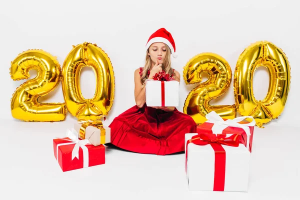 Pensive little girl w Santa kapelusz, stylowe dziecko siedzi zamyślony z prezentów świątecznych siedzi na białym tle ze złotem 2020 numery — Zdjęcie stockowe