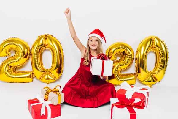 Lycklig liten flicka i Santa hatt, elegant baby jublar julklapp genom att visa seger gest medan du sitter på vit bakgrund med guld 2020 siffror — Stockfoto