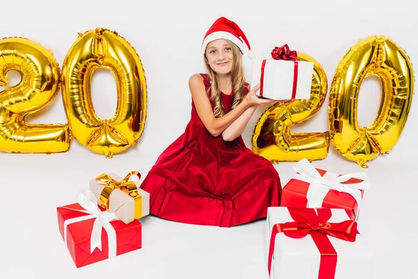 Glad liten flicka i Santa hatt, snygg baby sitter med julklappar sitter på vit bakgrund med guld 2020 siffror — Stockfoto