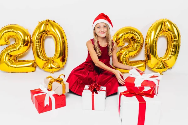 Glad liten flicka i Santa hatt, snygg baby sitter med julklappar sitter på vit bakgrund med guld 2020 siffror — Stockfoto