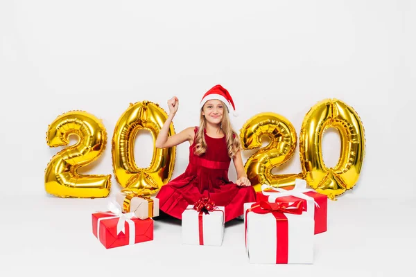 Glückliches kleines Mädchen mit Weihnachtsmütze, stilvolles Baby freut sich über Weihnachtsgeschenk, indem es Siegesgeste zeigt, während es auf weißem Hintergrund mit goldenen 2020-Zahlen sitzt — Stockfoto
