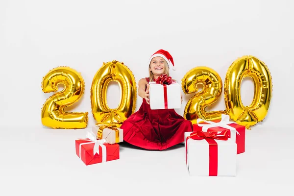 Joyeux petite fille dans le chapeau de Père Noël, élégant bébé donnant cadeau de Noël tout en regardant la caméra assis sur fond blanc avec des chiffres or 2020 — Photo