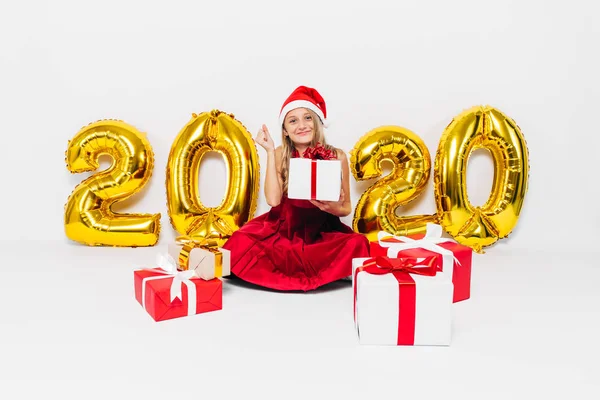Happy little girl w Santa kapelusz, stylowe dziecko raduje prezent świąteczny pokazując gest zwycięstwa siedząc na białym tle ze złotem 2020 numery — Zdjęcie stockowe