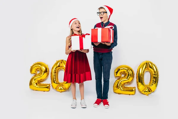 Menina e menino feliz vestindo chapéu de Papai Noel, irmão e irmã elegantes estão se alegrando segurando presentes de Natal, no fundo branco com figuras de ouro 2020 — Fotografia de Stock