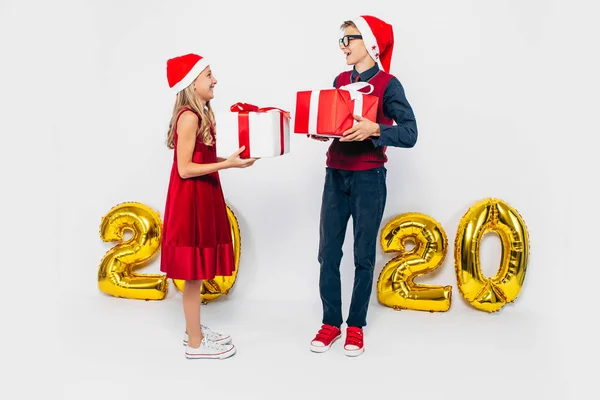 Szczęśliwa dziewczynka i chłopiec w kapeluszu Mikołaja, stylowy brat i siostra radują się i dają sobie prezenty, na białym tle ze złotymi figurami 2020 — Zdjęcie stockowe