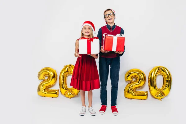 Menina e menino feliz vestindo chapéu de Papai Noel, irmão e irmã elegantes estão se alegrando segurando presentes de Natal, no fundo branco com figuras de ouro 2020 — Fotografia de Stock