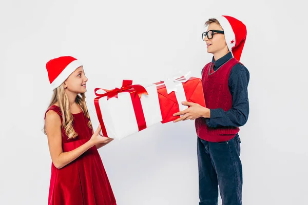 Szczęśliwa dziewczynka i chłopiec w Santa kapelusz, stylowy brat i siostra cieszą się i dając sobie prezenty, na białym tle — Zdjęcie stockowe