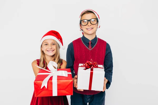 Szczęśliwa dziewczynka i chłopiec w Santa kapelusz, stylowy brat i siostra, szczęśliwie trzymając prezenty świąteczne w rękach, i patrząc na aparat stojący na białym tle — Zdjęcie stockowe