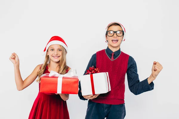 Szczęśliwa dziewczynka i chłopiec w kapeluszu Mikołaja, stylowy brat i siostra, radują się trzymając prezenty świąteczne w rękach, i pokazać gest zwycięstwa stojąc na białym tle — Zdjęcie stockowe