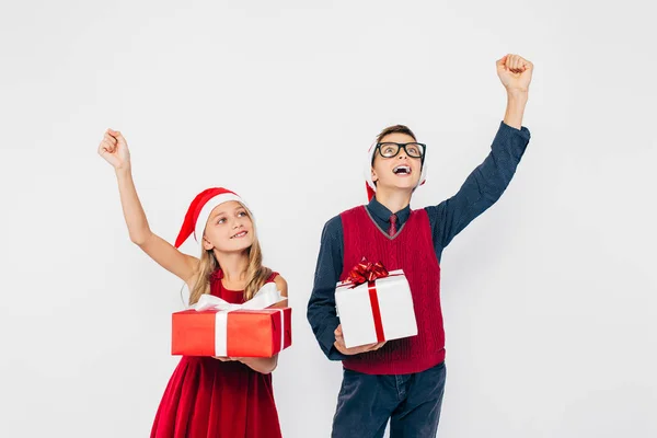 Szczęśliwa dziewczynka i chłopiec w kapeluszu Mikołaja, stylowy brat i siostra, radują się trzymając prezenty świąteczne w rękach, i pokazać gest zwycięstwa stojąc na białym tle — Zdjęcie stockowe