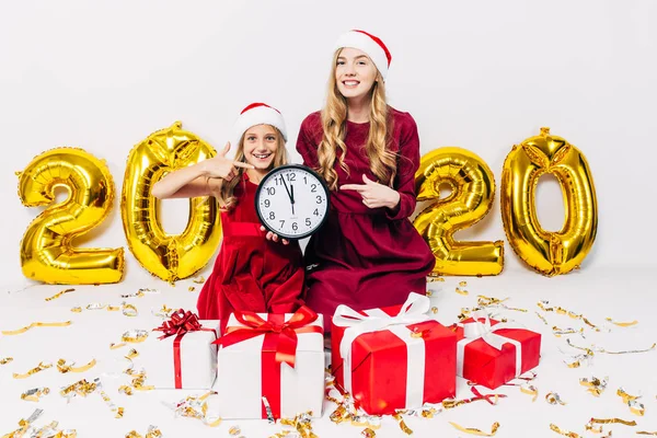 Jovem família de Natal, mãe e filhinha em chapéus de Papai Noel, com relógio de parede na mão e apontando o dedo para eles, sentado com presentes de Natal, no fundo branco — Fotografia de Stock