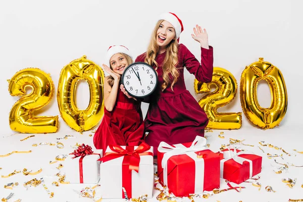 Família de Natal, mãe e filhinha em chapéus de Papai Noel, se divertindo mostrando um gesto de vitória segurando um relógio de parede enquanto se senta com presentes de Natal — Fotografia de Stock