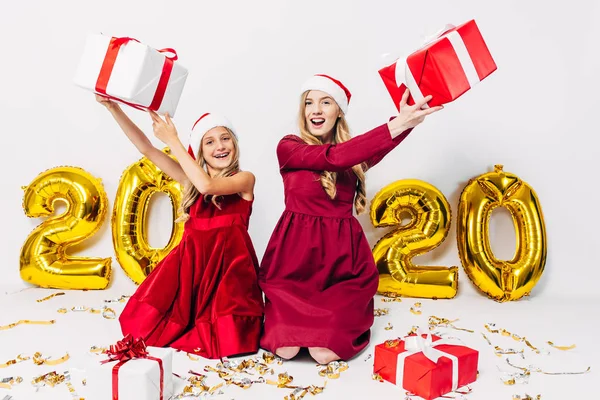 Eine glückliche Mutter und kleine Tochter in Weihnachtsmannmützen, die mit Weihnachtsgeschenken in der Hand auf einem weißen Hintergrund sitzen und Spaß haben — Stockfoto