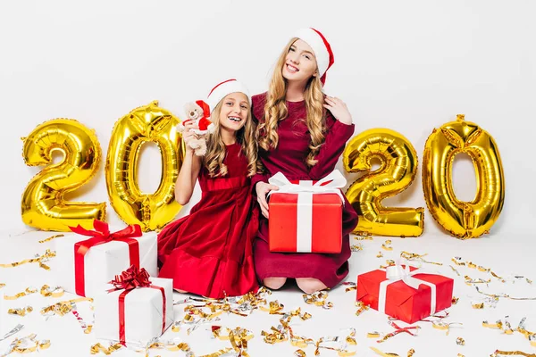 Szczęśliwa matka i córeczka w kapeluszach Mikołaja, bawiące się siedząc na białym tle z prezentami świątecznymi w rękach — Zdjęcie stockowe