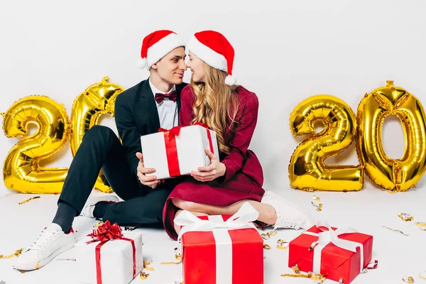 Jovem família de Natal, cara e menina em chapéus de Papai Noel abraçando uns aos outros e segurando presentes de Natal enquanto sentado no fundo branco — Fotografia de Stock