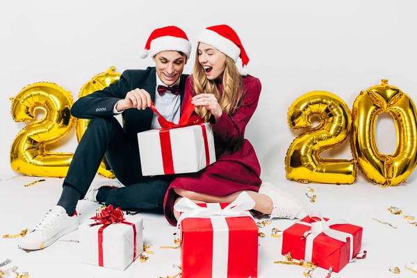 Młoda świąteczna rodzina, chłopak i dziewczyna w kapeluszach Mikołaja otwierają prezenty świąteczne siedząc na białym tle — Zdjęcie stockowe