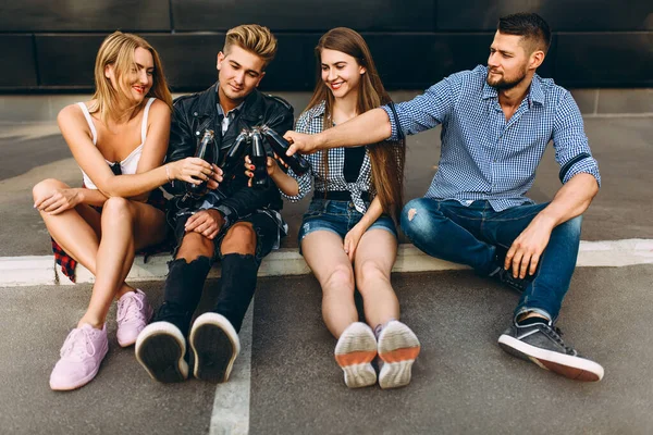Grupa stylowych szczęśliwych przyjaciół, dziewczyn i facetów siedzi razem w mieście na asfalcie przy czarnej ścianie, bawiąc się i pijąc drinki — Zdjęcie stockowe