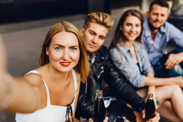 Группа веселых друзей, парней и девушек гуляют по городу, делая селфи на смартфоне, веселятся и пьют напитки — стоковое фото