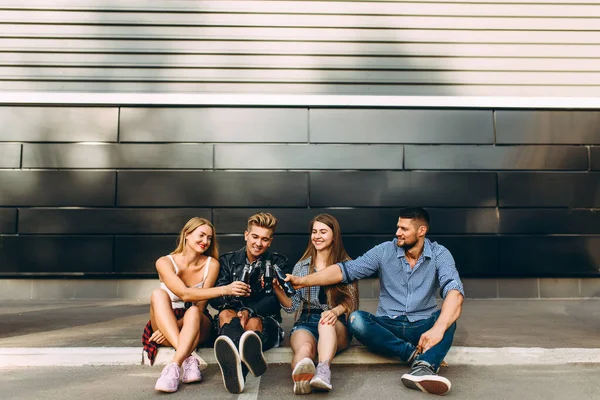 Un grupo de elegantes amigos felices, chicas y chicos se sientan juntos en la ciudad en el asfalto en la pared negra, divirtiéndose y bebiendo bebidas —  Fotos de Stock