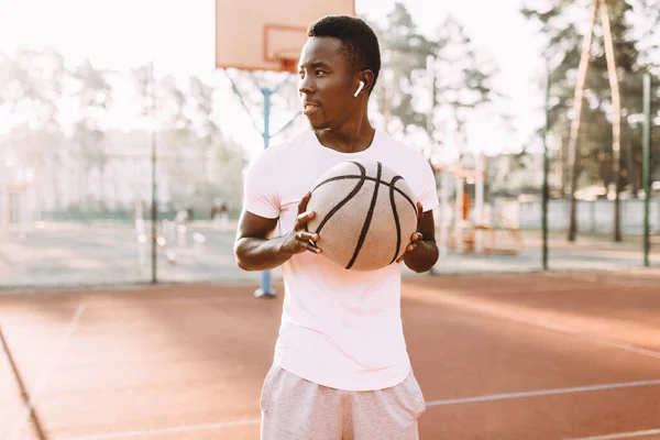 Sportiv tânăr afro-american în picioare pe teren în zori cu o minge de baschet — Fotografie, imagine de stoc