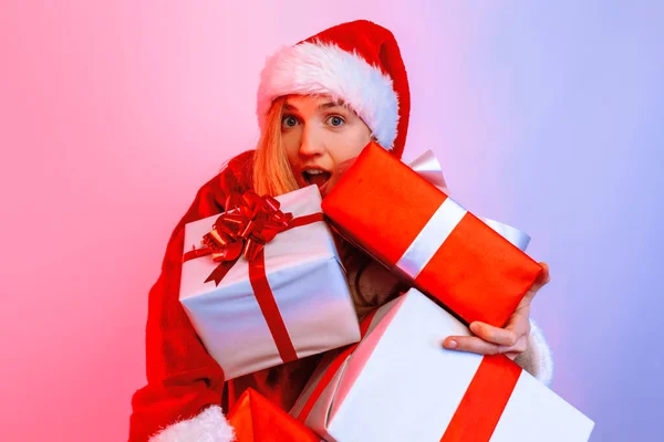 Uma loira surpreso em um chapéu de Papai Noel espreita para fora de uma pilha de presentes em pé no fundo — Fotografia de Stock