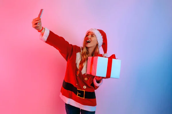 Happy young woman in Santa hat taking selfie on smartphone standing on background — Stock Photo, Image
